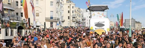 Crowds of people gathering on Marine Parade during a sunny outdoor event, with rainbow flags and buildings in the background.