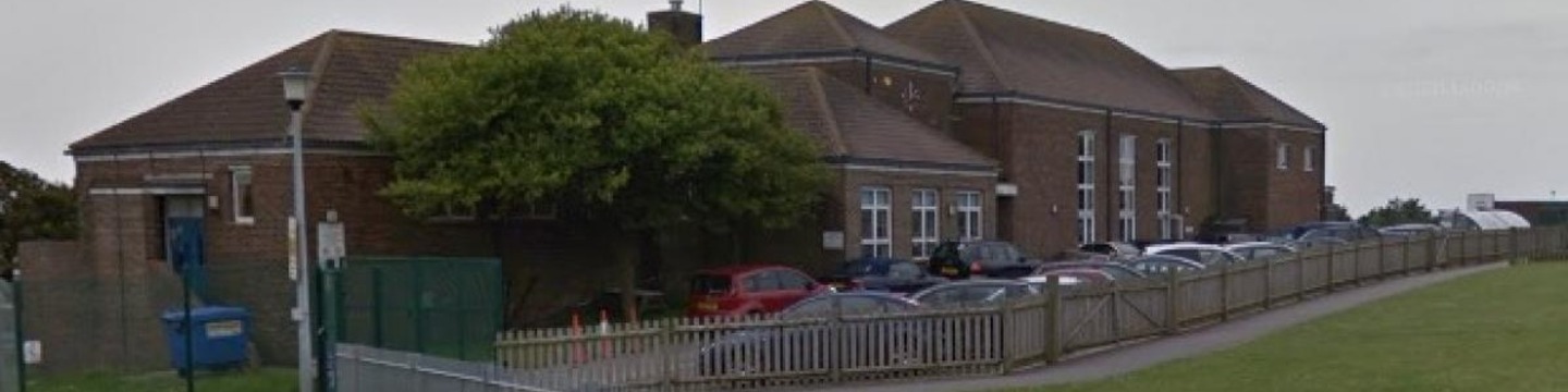 An image showing Woodingdean Primary School. A grassy field is in the foreground and a number of cars are parked in front of the school building.