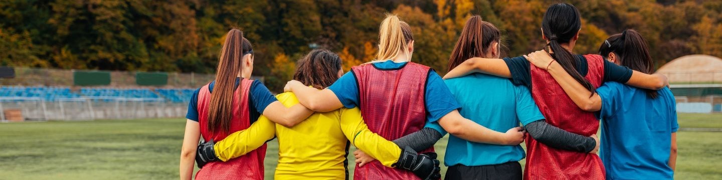 Six women in sports bibs facing away from the camera with their arms around each others backs