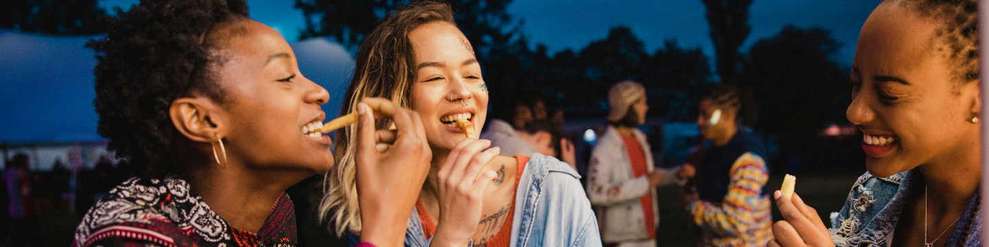 Three friends laughing as they enjoy a cone of chips at a night time gathering