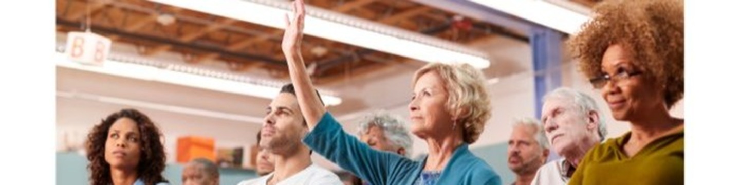The image shows a group of people sitting down with one person with their hand raised to ask a question