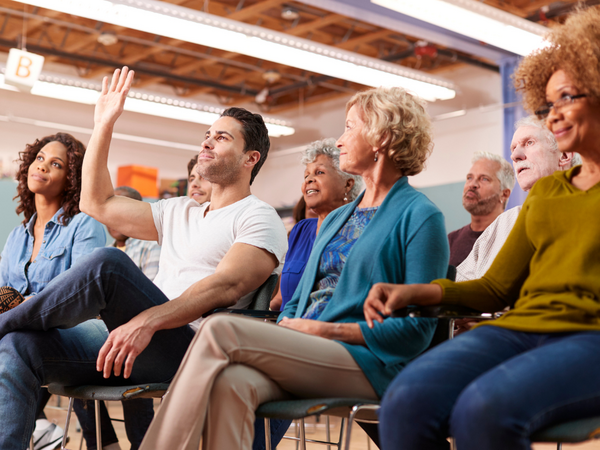 The image shows a group of people sitting down with one person with their hand raised to ask a question.