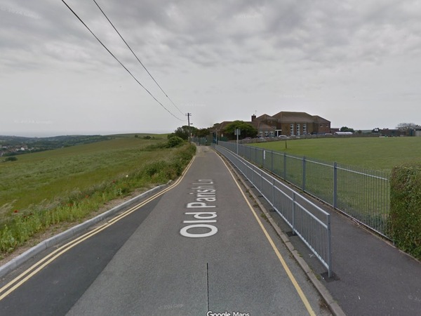A map showing Old Parish Lane looking towards Woodingdean Primary School. A field enclosed by a fence is on the right side of the image and open fields are to the left. There are double yellow lines along the road on the left side and single yellow line along the right side of the road. A barrier is on the pavement on the right side of the road. The school is in centre of the image and power lines are in the centre of the top of the image.